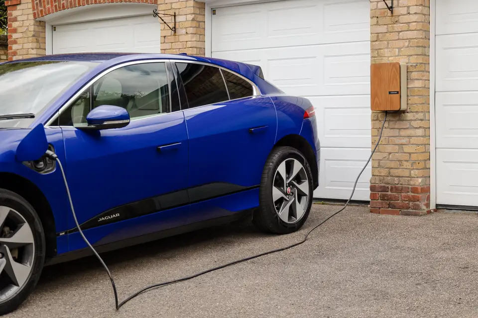 A blue electric car charges from a wall box beside a row of garage doors.